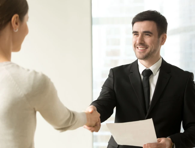 Hombre de traje felicitando a una mujer con un apretón de manos mientras le entrega un documento, en una oficina luminosa Hombre de traje felicitando a una mujer con un apretón de manos mientras le entrega un documento, en una oficina luminosa