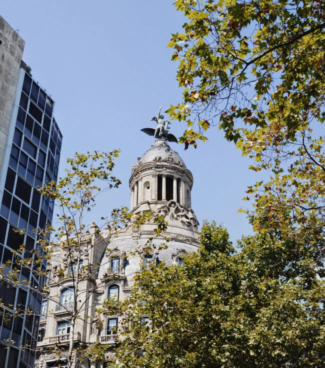 Fachada de edificio histórico con cúpula y escultura en lo alto, rodeado de árboles en una calle de Barcelona Fachada de edificio histórico con cúpula y escultura en lo alto, rodeado de árboles en una calle de Barcelona