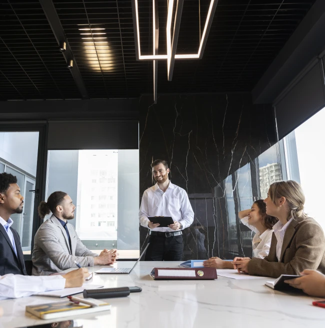 Reunión de negocios en una sala moderna, con un hombre de pie presentando mientras un grupo diverso de personas lo escucha atentamente alrededor de una mesa de reuniones Reunión de negocios en una sala moderna, con un hombre de pie presentando mientras un grupo diverso de personas lo escucha atentamente alrededor de una mesa de reuniones
