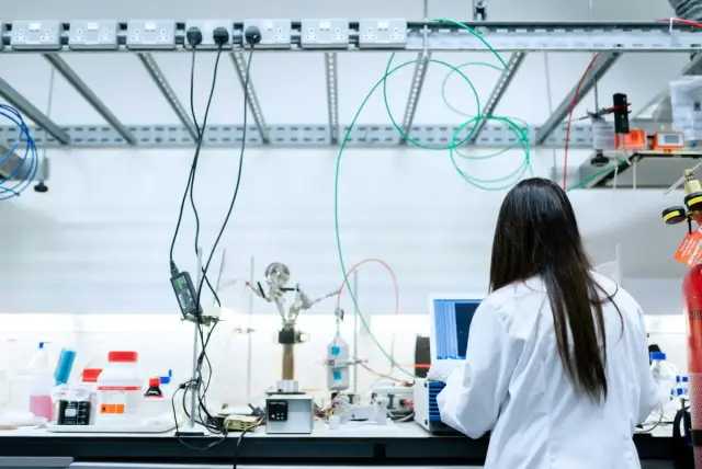 Científica con bata blanca trabajando de espaldas con equipos en un laboratorio