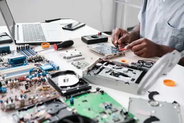Técnico reparando componentes electrónicos sobre una mesa con placas y un portátil