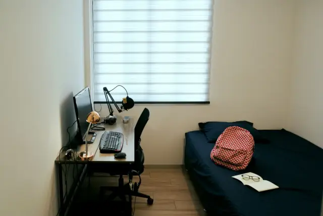 Habitación individual con escritorio, ordenador, cama y una mochila a cuadros